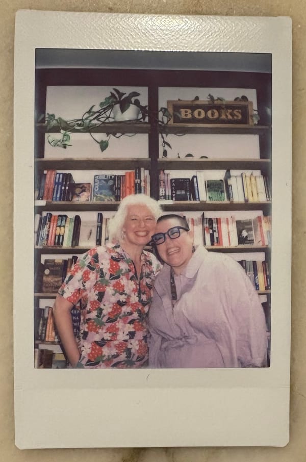 A photograph of a polaroid picture taken at Windowseat Books in Nanaimo, BC. Zena and Hannah are standing side-by-side in front of a bookshelf, both grinning broadly.